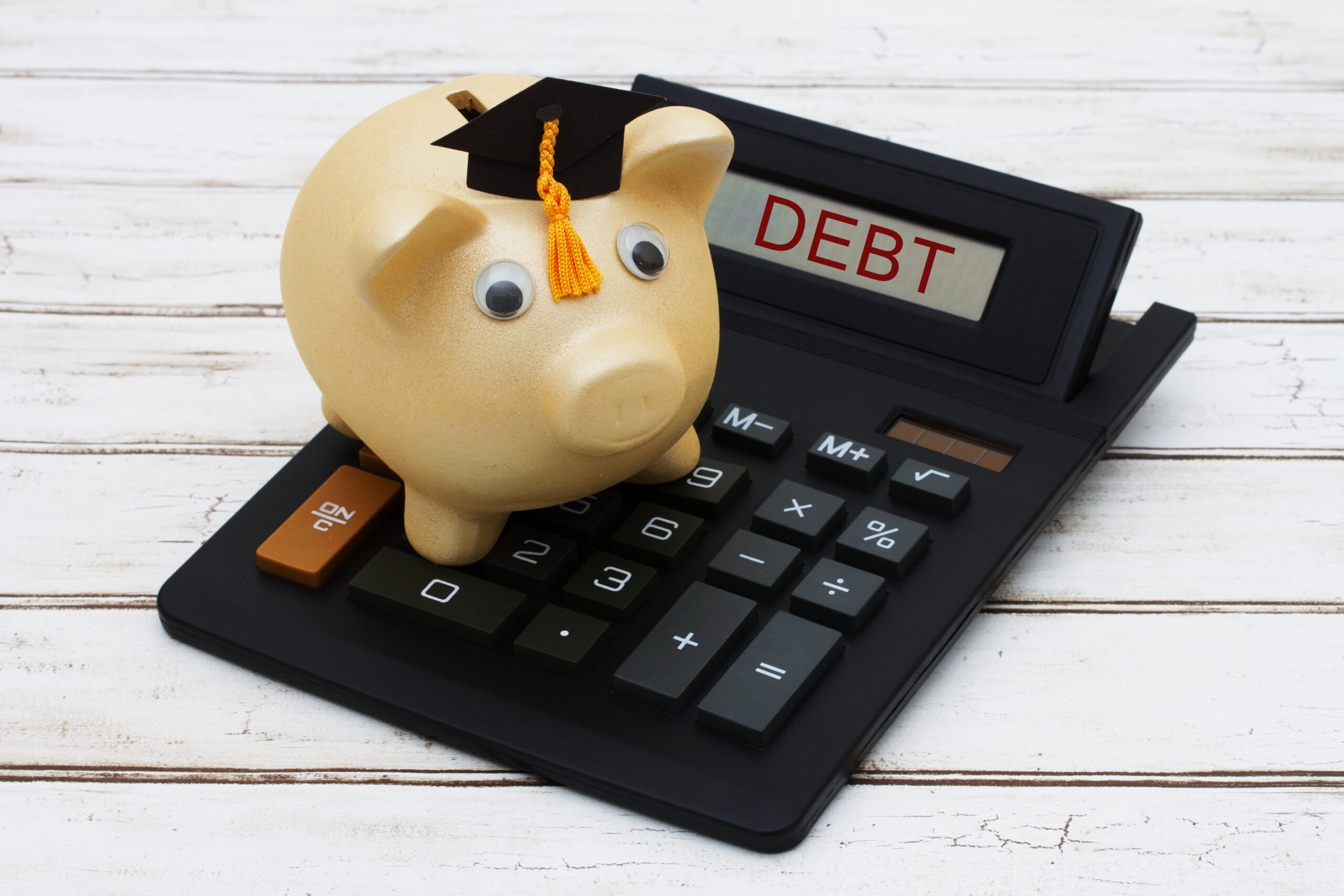 Calculating your student loan debt, A golden piggy bank with a grad hat on a calculator with word Debt over a distressed wood background