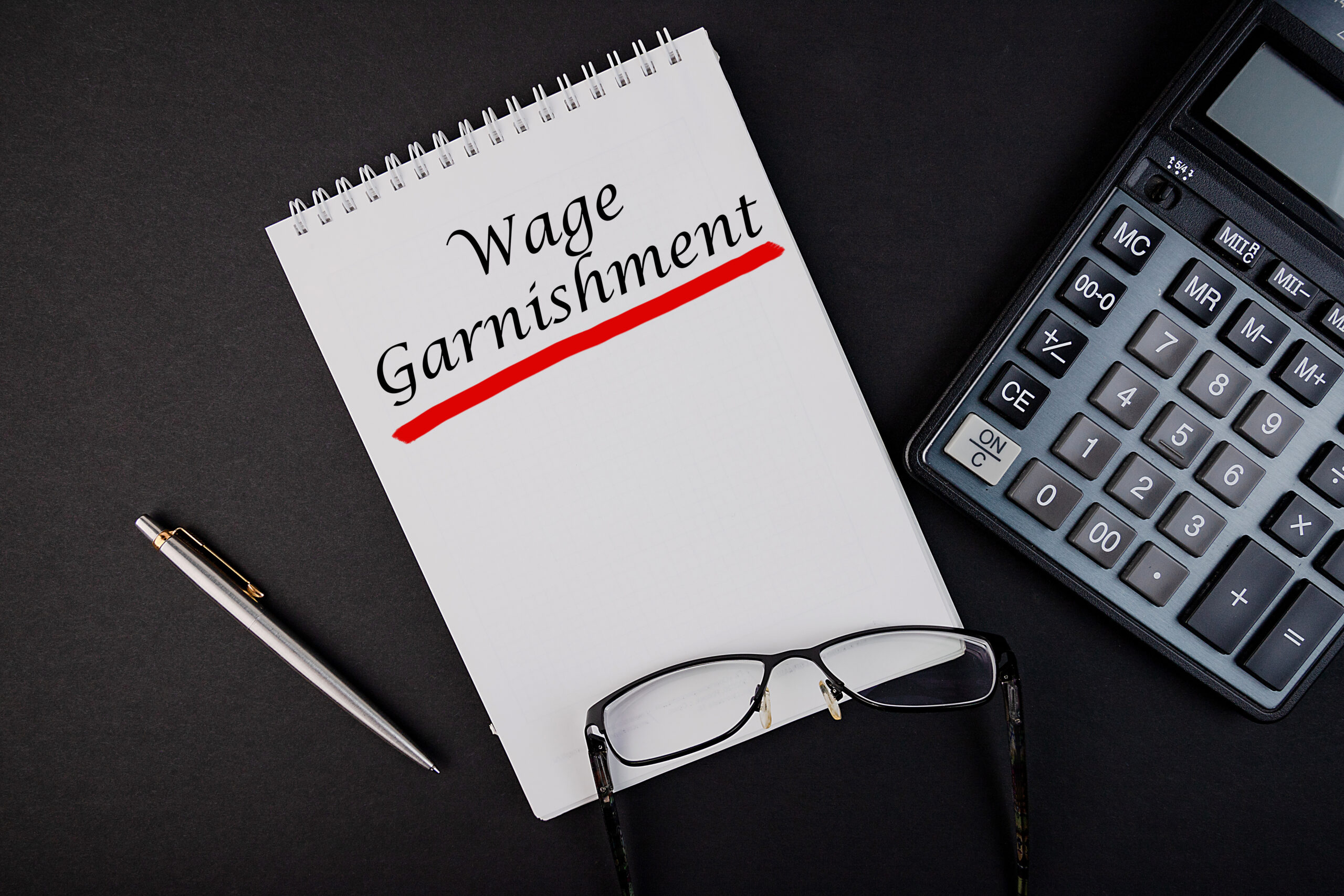 Wage Garnishment word concept written in a notebook with pen and calculator, top view.
